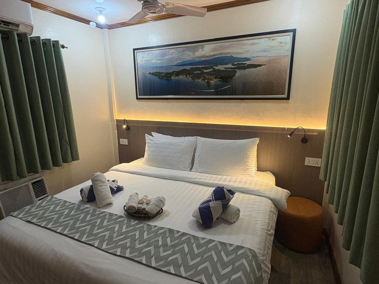 A neatly made bed with white linens and blue and white towels, a headboard, a wall-mounted light, and a painting of a landscape.