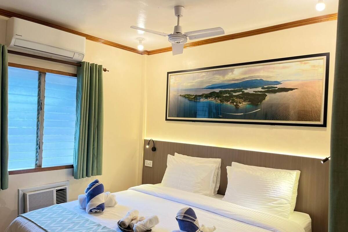 A hotel room with a bed, a ceiling fan, an air conditioning unit, a window with curtains, and a painting on the wall.