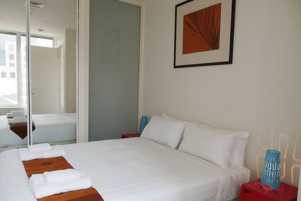 A hotel room with a large bed, a red nightstand, a blue vase, and a framed picture on the wall.