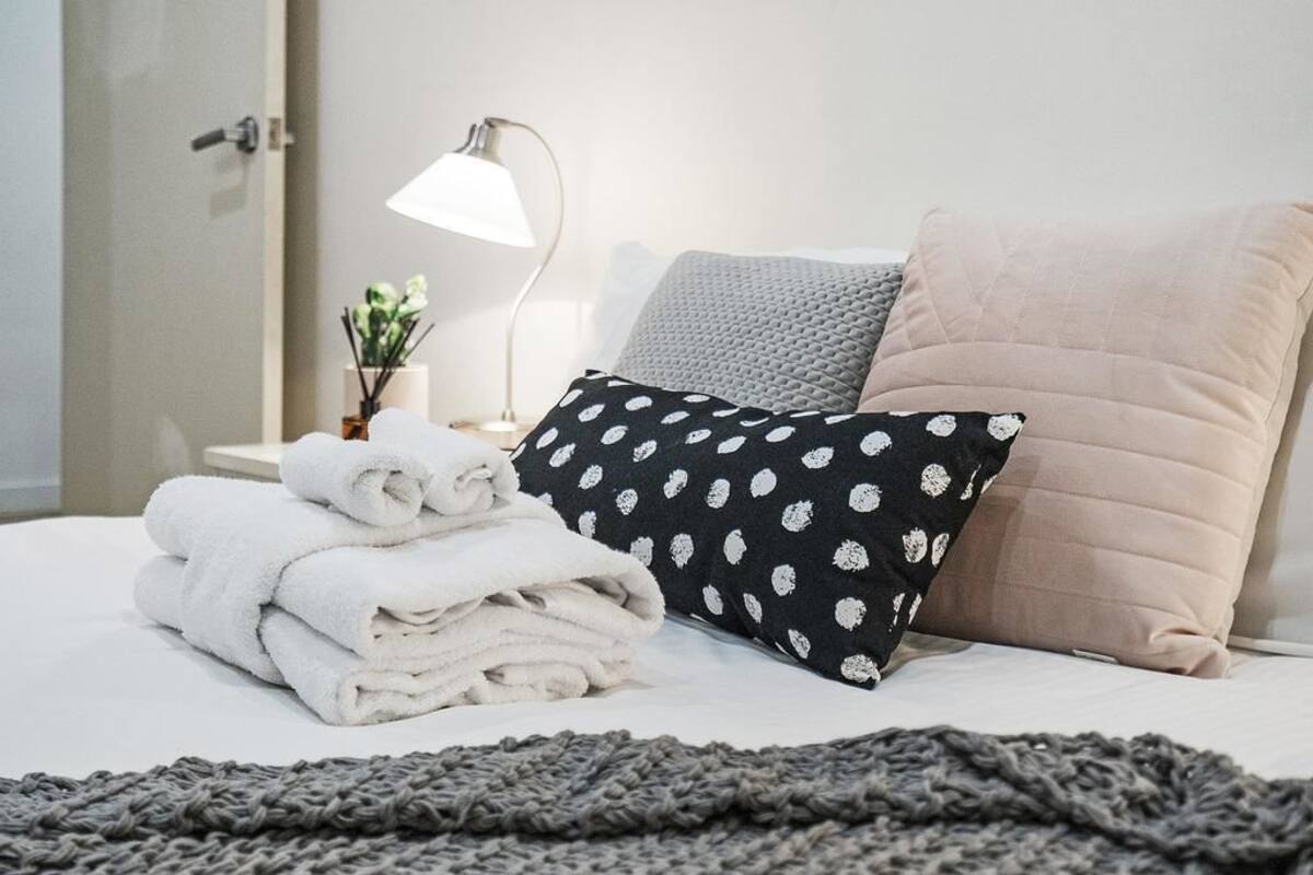 A neatly made bed with folded white towels, a black pillow with white floral patterns, a beige pillow, and a gray knitted blanket.