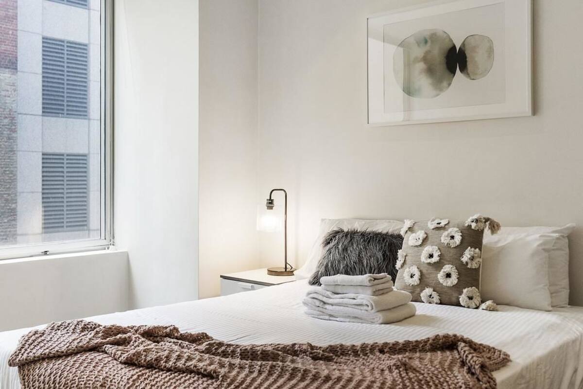 A neatly made bed with a blanket, pillows, and folded towels. There is a framed artwork on the wall and a bedside lamp.