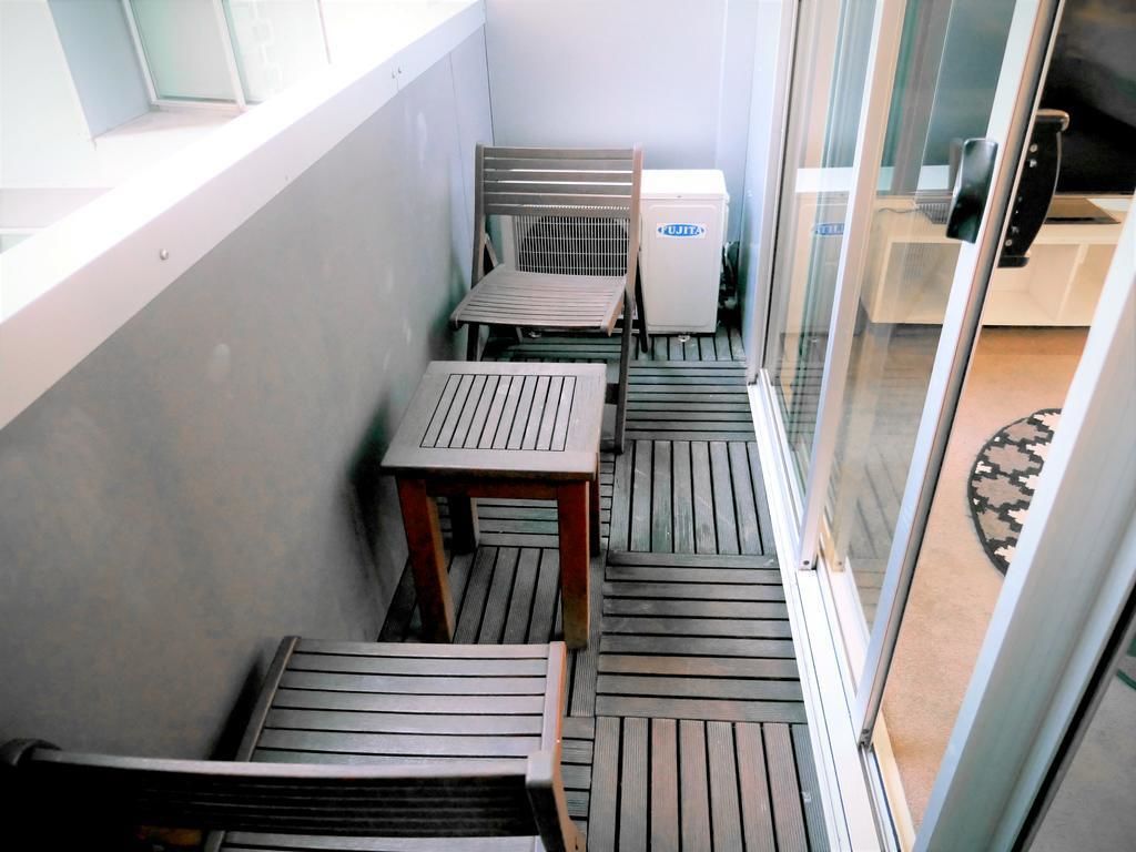 A balcony with wooden tables and chairs, a small refrigerator, and a glass door leading to a room.