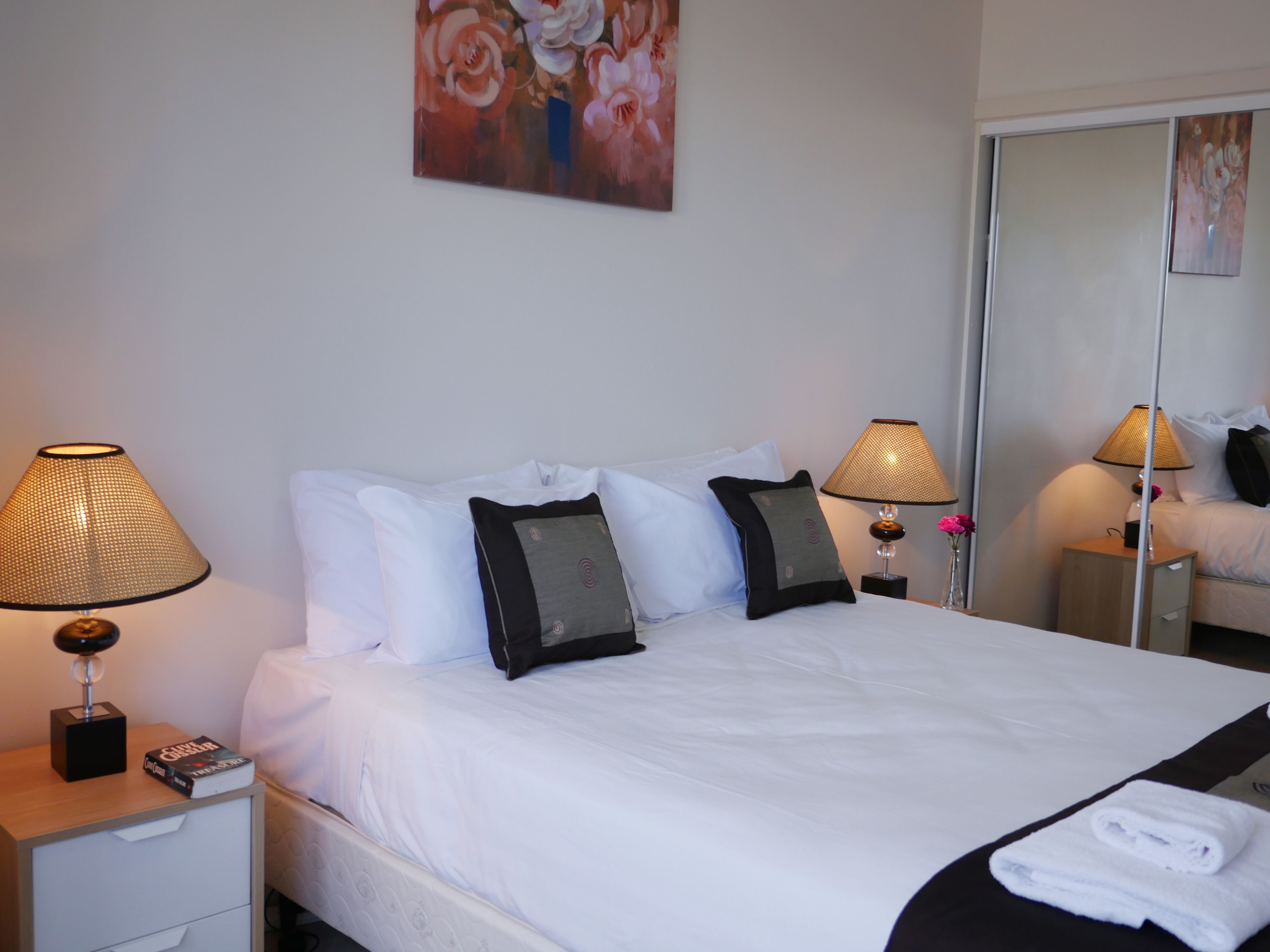 A hotel room with a neatly made bed, two bedside lamps, a nightstand with a book, and a mirror reflecting the room.