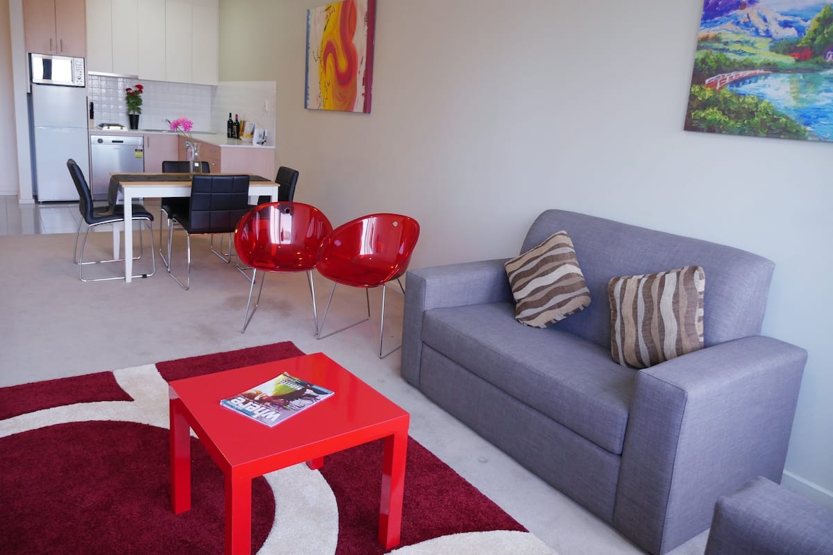 A modern living room with a grey sofa, red chairs, and a red table. A kitchen is visible in the background with white cabinets and a microwave.