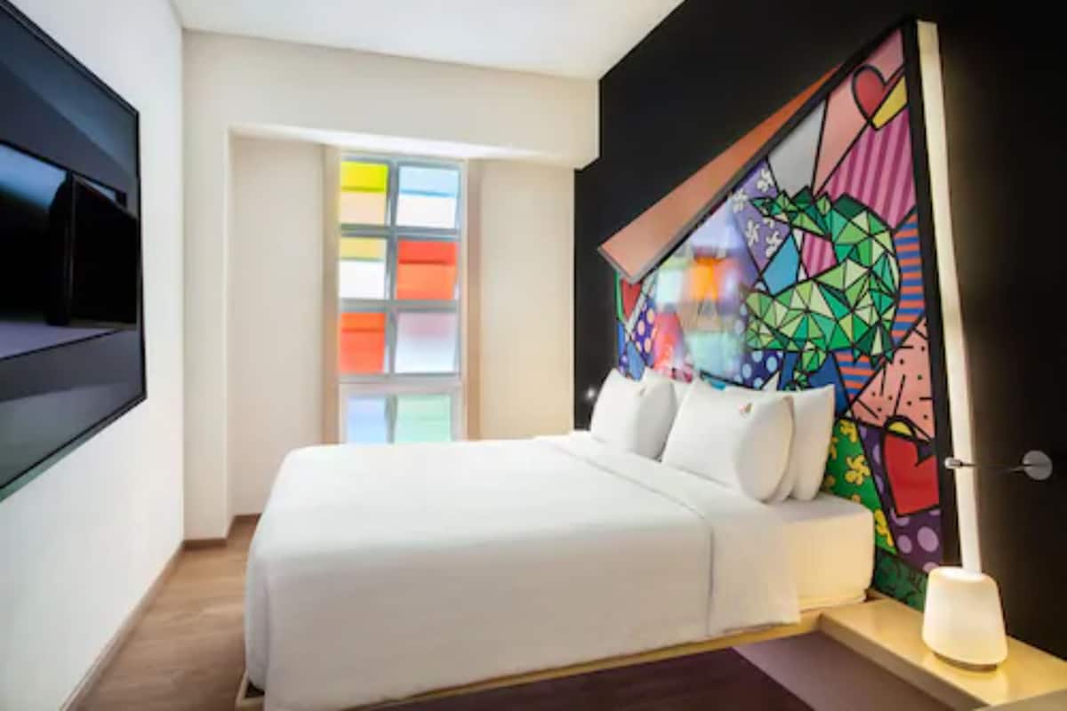 A modern hotel room with a large bed, a flat-screen TV mounted on the wall, and a colorful geometric artwork above the bed.