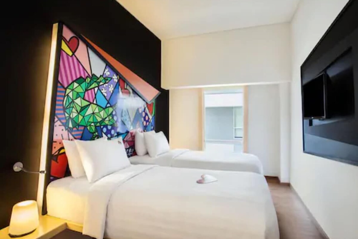 Habitación de hotel con dos camas, un cuadro abstracto grande en la pared, un televisor de pantalla plana montado y una ventana que deja entrar luz natural.