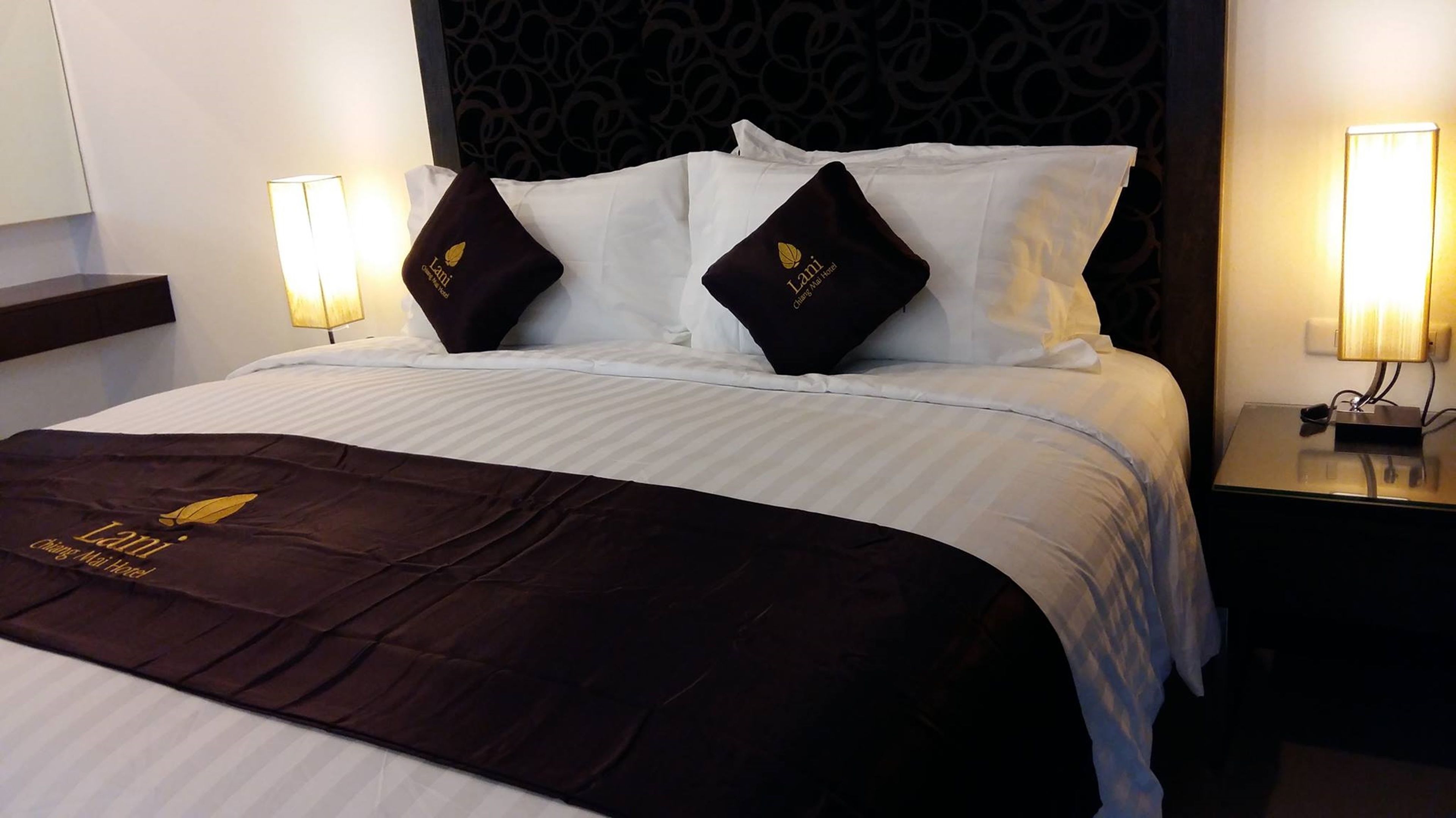 A hotel room with a neatly made bed, two pillows with a logo, a dark brown bedspread, and bedside lamps.