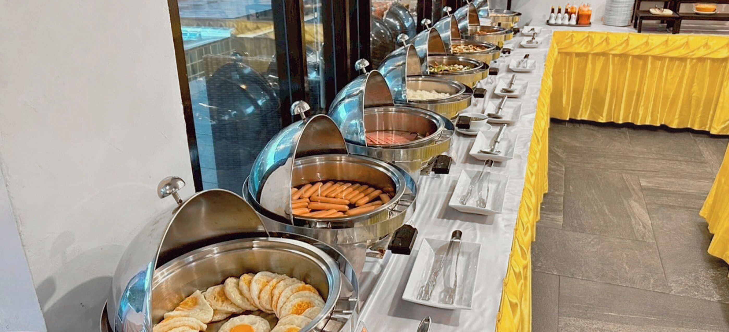 A buffet setup with various food items in metal containers, arranged on a table with white plates and cutlery.