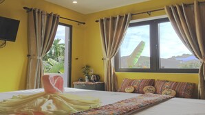 A room with yellow walls, a bed with a patterned bedspread, a small table with a plant, and a window with curtains. - Longan Homestay (Da Nang)