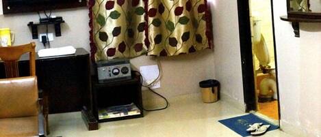 Une pièce avec un téléviseur fixé au mur, une chaise en bois, une petite table, un radiateur et un rideau orné d’un motif de feuilles.