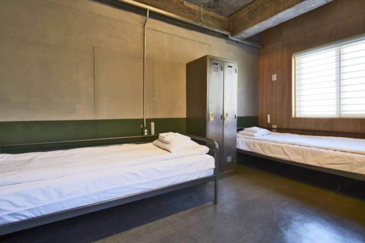 Habitación tipo dormitorio con dos camas individuales, un armario y una ventana con persianas.