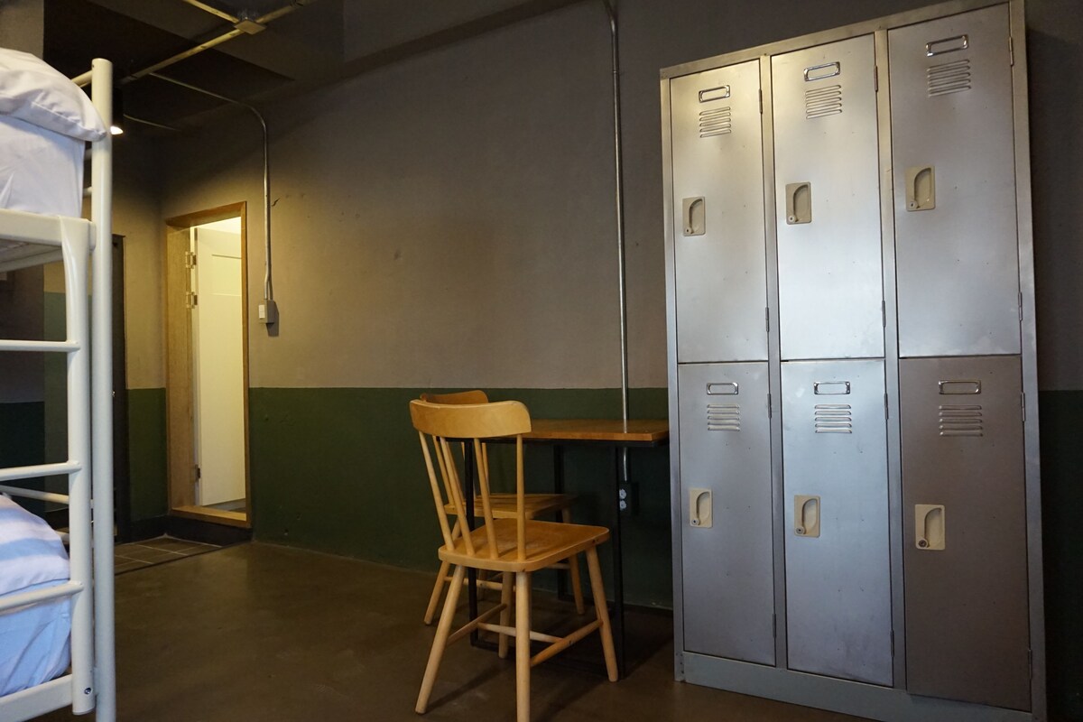 A dormitory room with bunk beds, a wooden chair, a table, and metal lockers.
