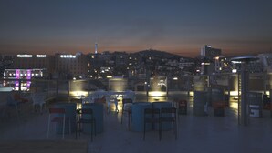 Una terraza en la azotea con sillas y mesas, vistas de la ciudad y un edificio con señalización iluminada.