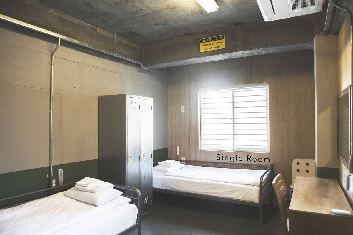 Une chambre individuelle avec deux lits, un bureau, une chaise, une armoire et une fenêtre avec des stores.