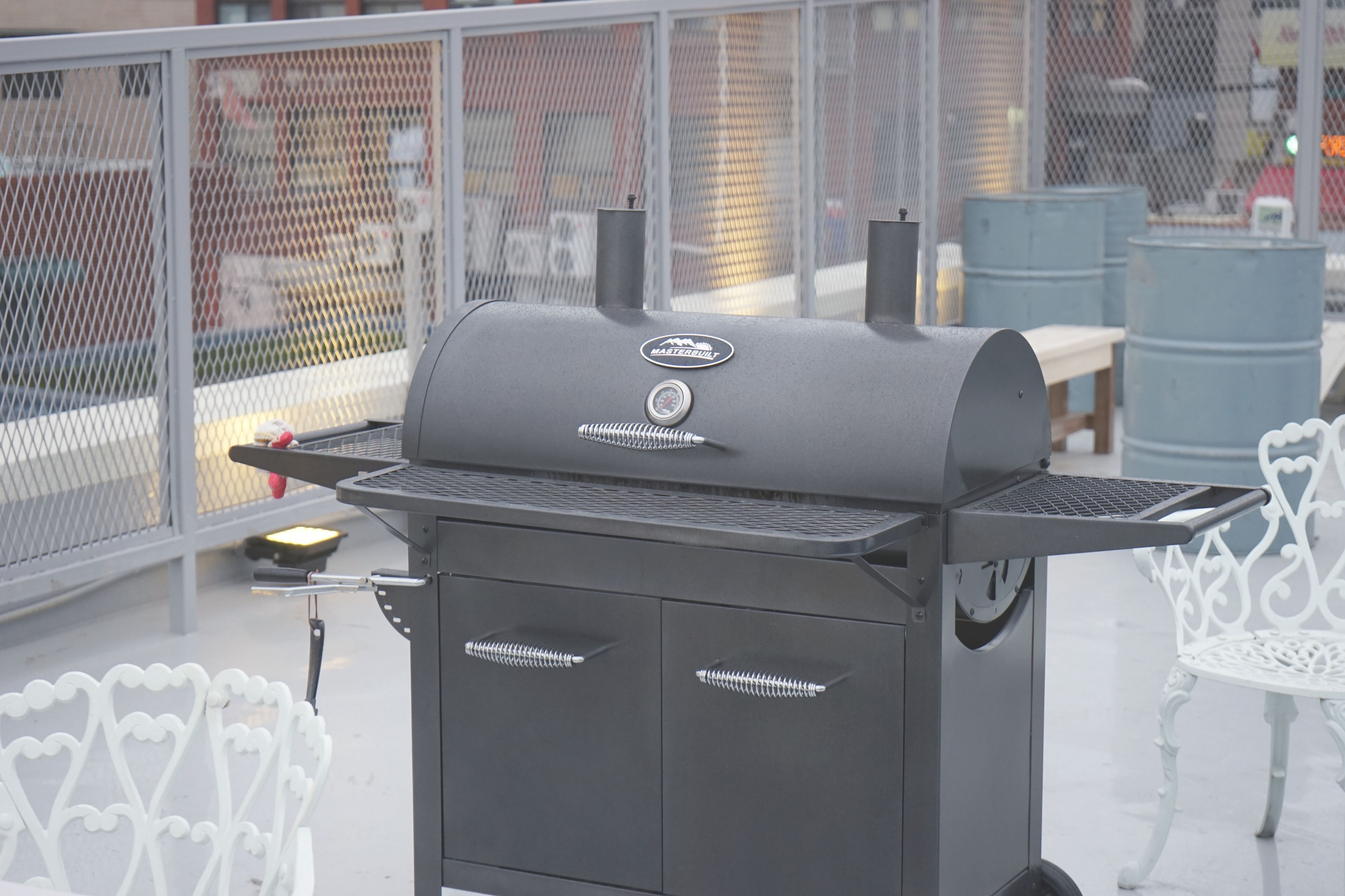 Una parrilla para barbacoa con panel de control y logotipo, ubicada al aire libre con una valla metálica y sillas blancas al fondo.