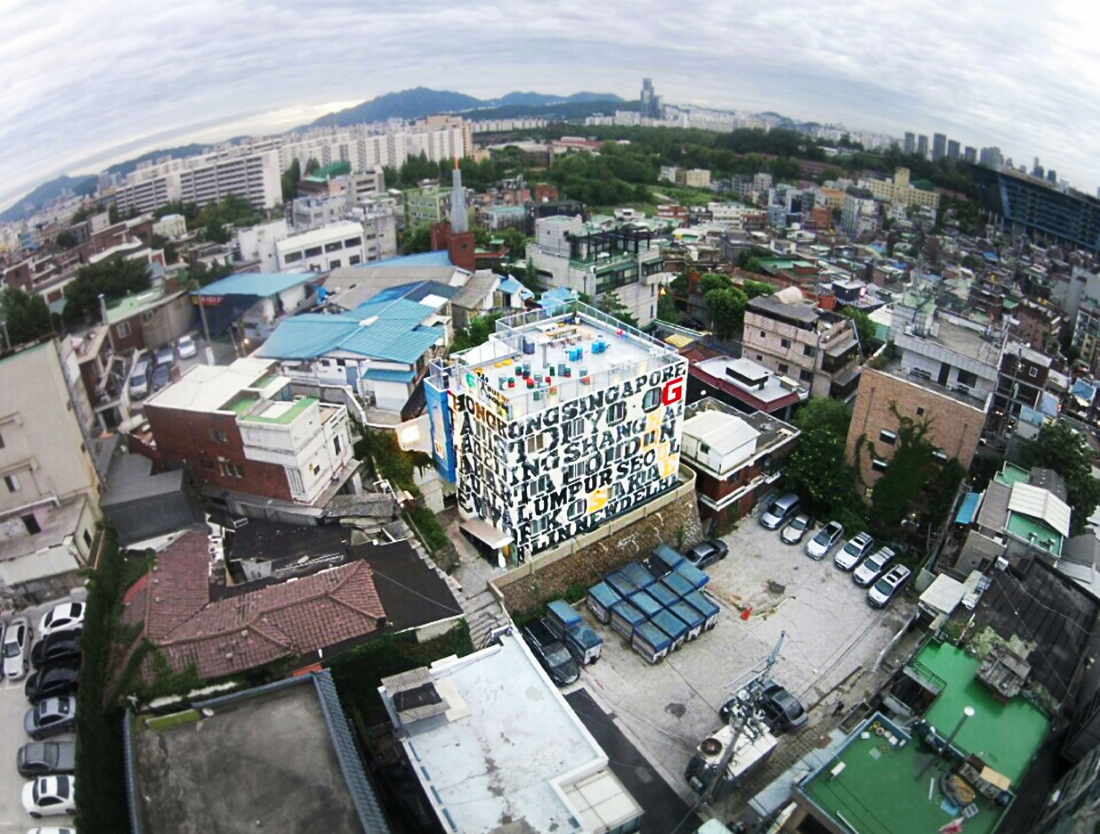 Vista aérea de uma área urbana densamente construída, com diversos edifícios, incluindo uma estrutura imponente de vários andares com um telhado coberto de letras e símbolos coloridos.