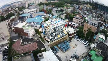 Vista aérea de uma área urbana densamente construída, com diversos edifícios, incluindo uma estrutura imponente de vários andares com um telhado coberto de letras e símbolos coloridos.