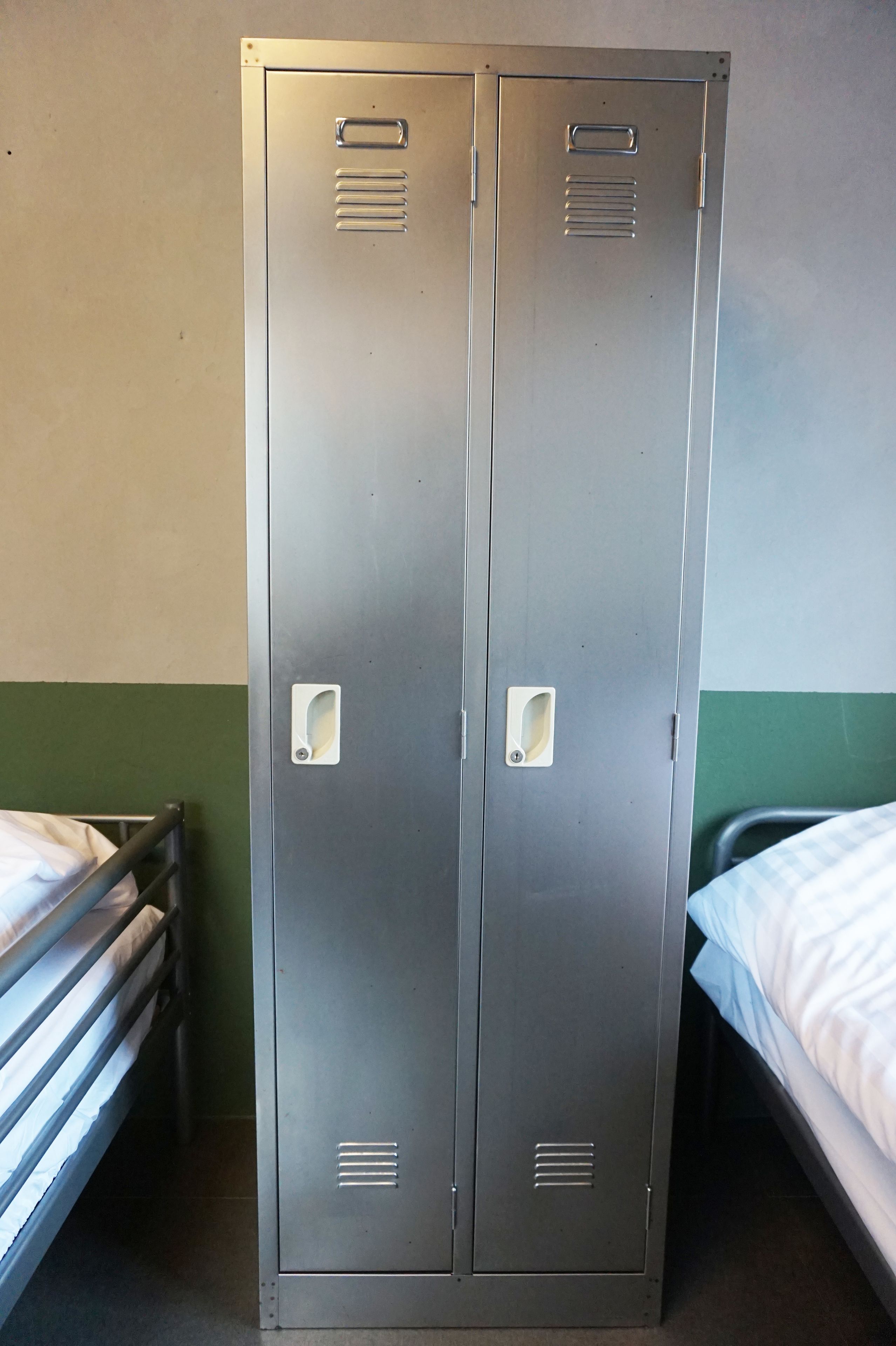Un vestuario metálico con dos taquillas, una cama con ropa blanca y una pared verde.