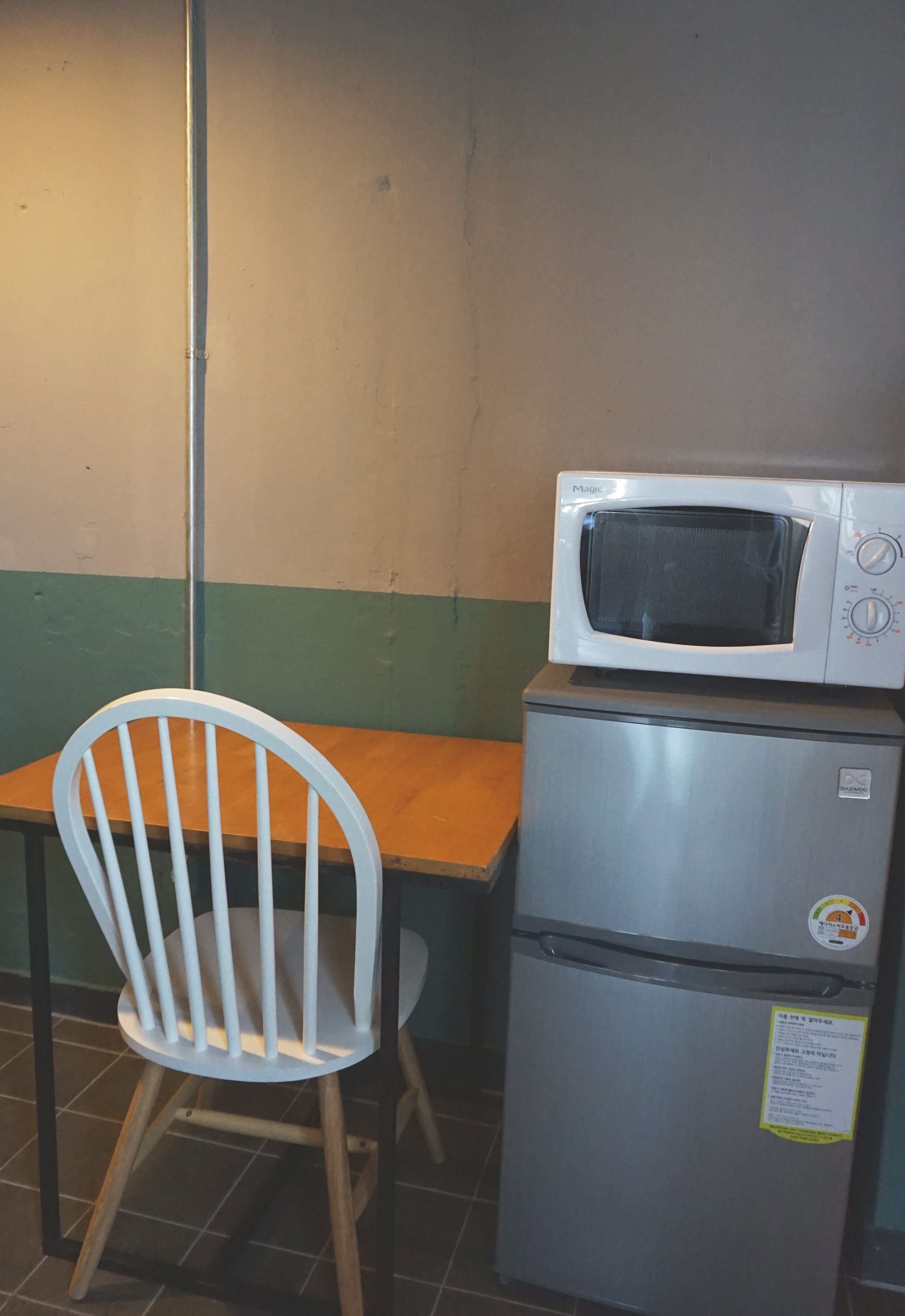 A small kitchenette with a microwave on top of a refrigerator, a wooden chair, and a tiled floor.