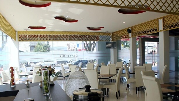 A modern restaurant interior with white chairs, a long table with a vase of flowers, and a view of the street outside. - Idea's Hotel (Bandung)