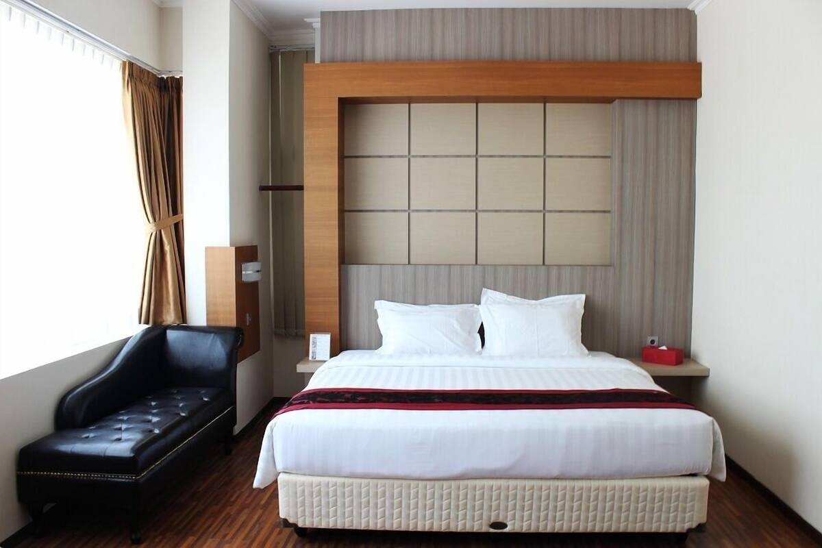 A hotel room with a large bed, a black leather sofa, a wooden headboard, and a window with curtains.