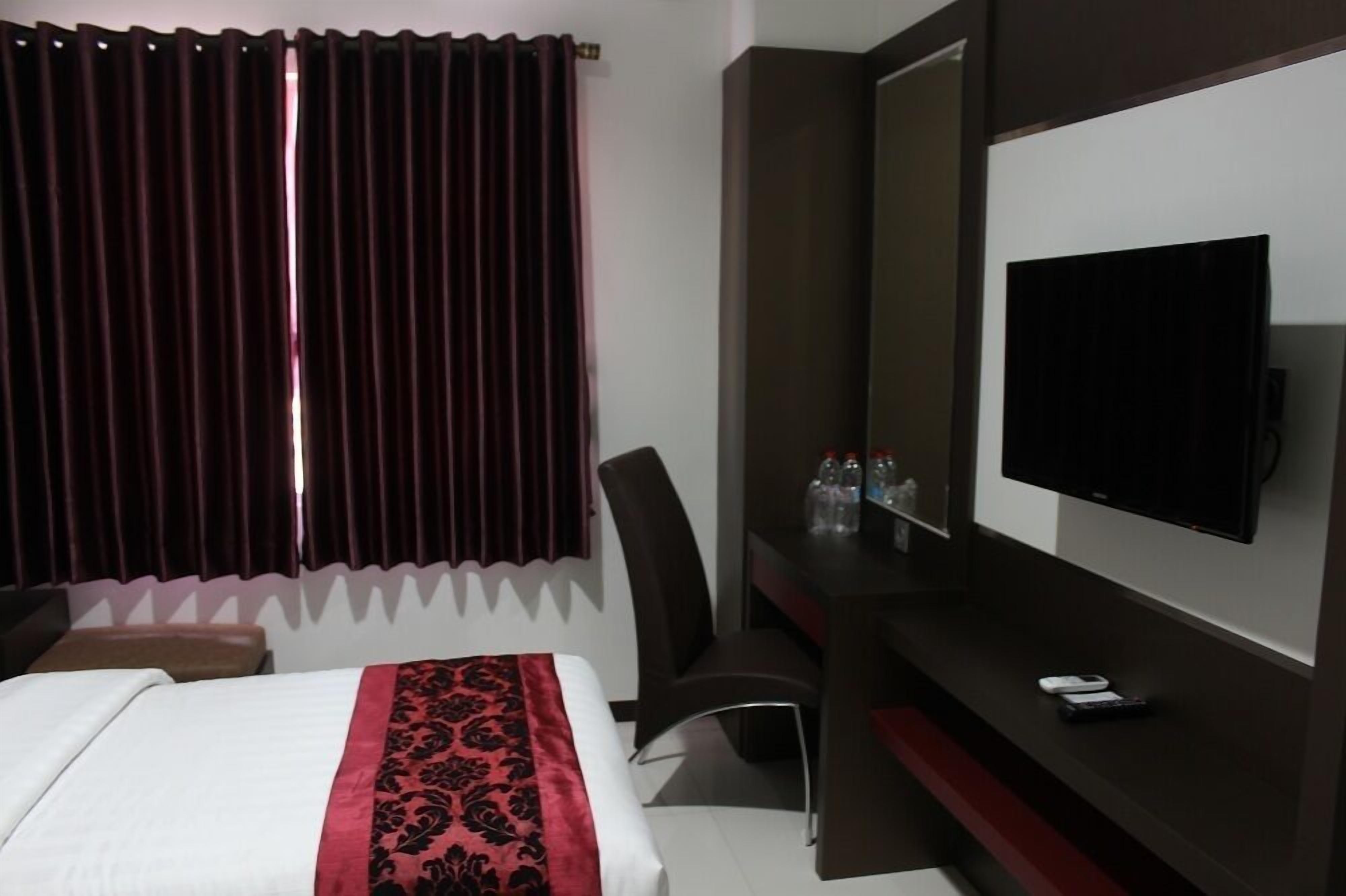 A hotel room with a bed, a desk with a TV, a chair, and a window with curtains.