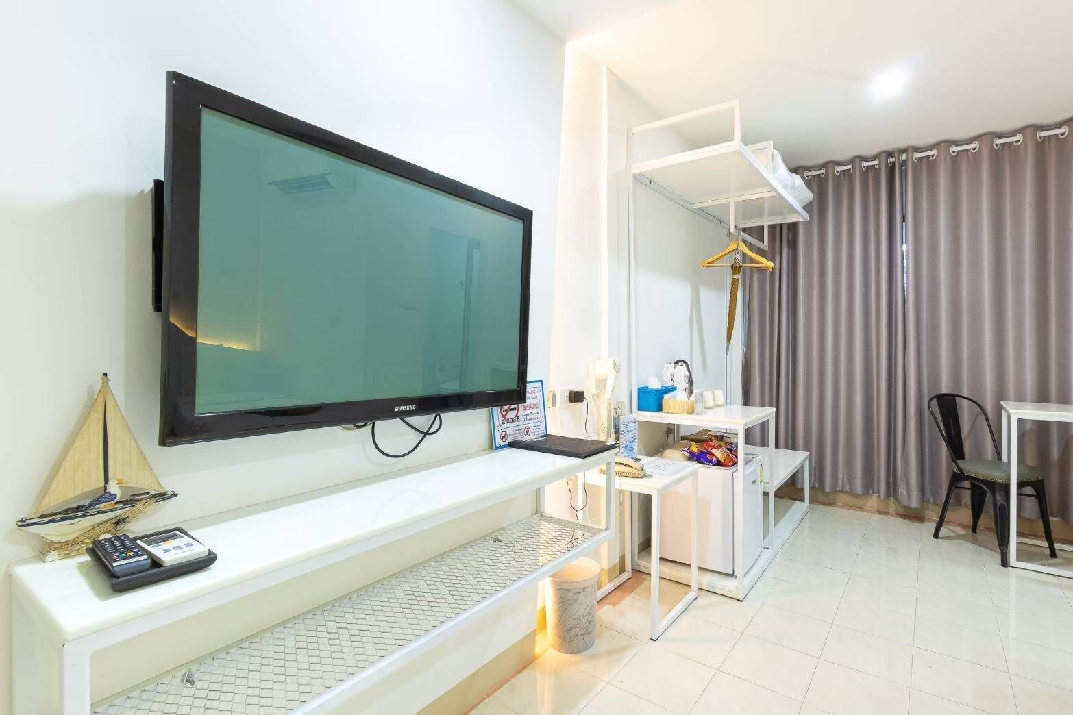 Habitación de hotel moderna con una televisión de pantalla plana grande montada en la pared, un escritorio pequeño con una computadora portátil, una silla y un perchero.