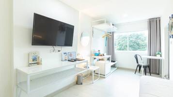 A hotel room with a flat-screen TV, a desk with a chair, a bed, a window with curtains, and a small plant on the bedside table.