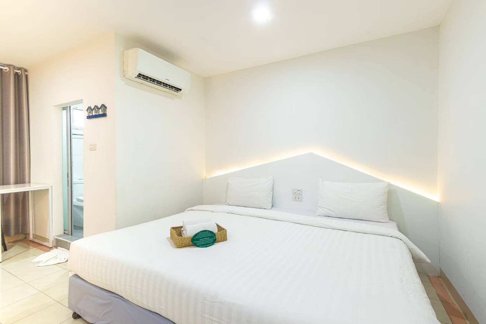 Un lit bien fait, avec du linge de lit et des oreillers blancs, un petit panier en osier contenant un objet vert, et un climatiseur fixé au mur.
