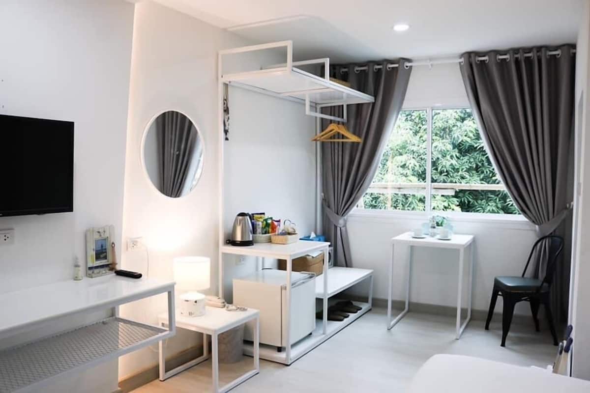 A modern hotel room with a desk, a small table, a chair, a TV, a window with curtains, and a shelf unit.