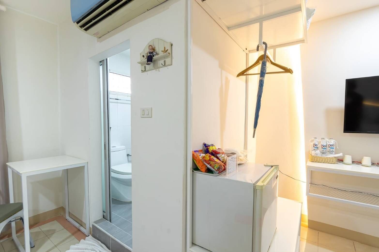 Habitación de hotel con una decoración en tonos blancos, una pequeña mesa, una silla, perchero, televisor y un minibar con aperitivos y bebidas.