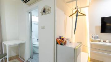 Habitación de hotel con una decoración en tonos blancos, una pequeña mesa, una silla, perchero, televisor y un minibar con aperitivos y bebidas.