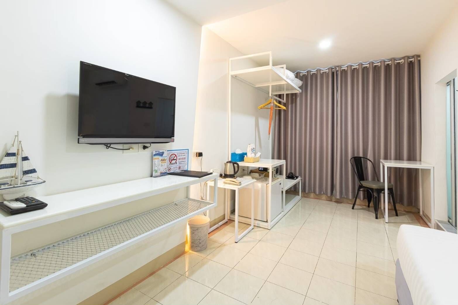 A modern hotel room with a flat-screen TV, a desk with a chair, a small table with a kettle, a coat rack, and a bed.