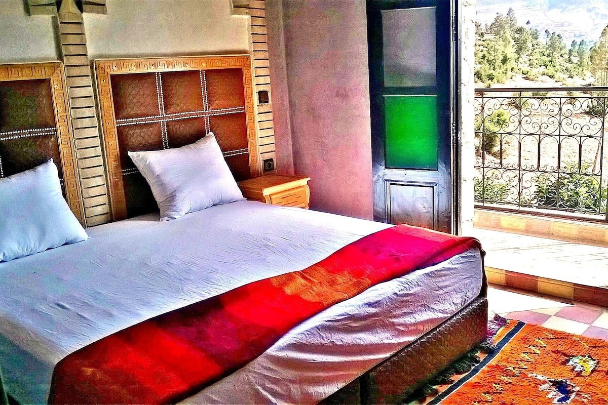 A hotel room with a large bed, a wooden headboard, a nightstand, a TV, and a balcony with a view of trees and mountains.