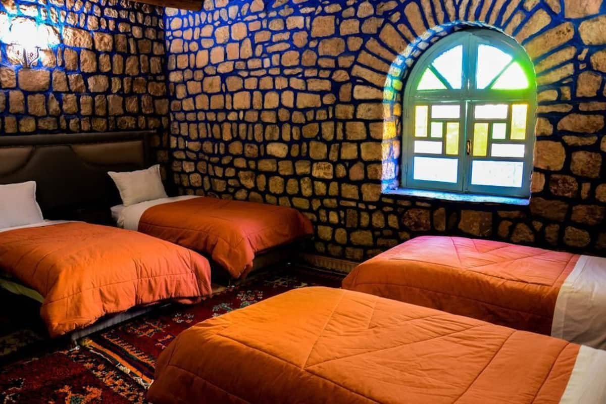 A room with stone walls, a large arched window, and three beds with orange bedding.