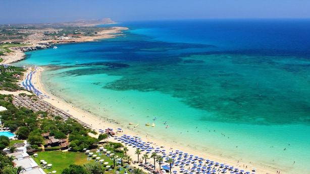 Uma praia costeira com águas turquesa cristalinas, areal arenoso e diversos guarda-sóis.