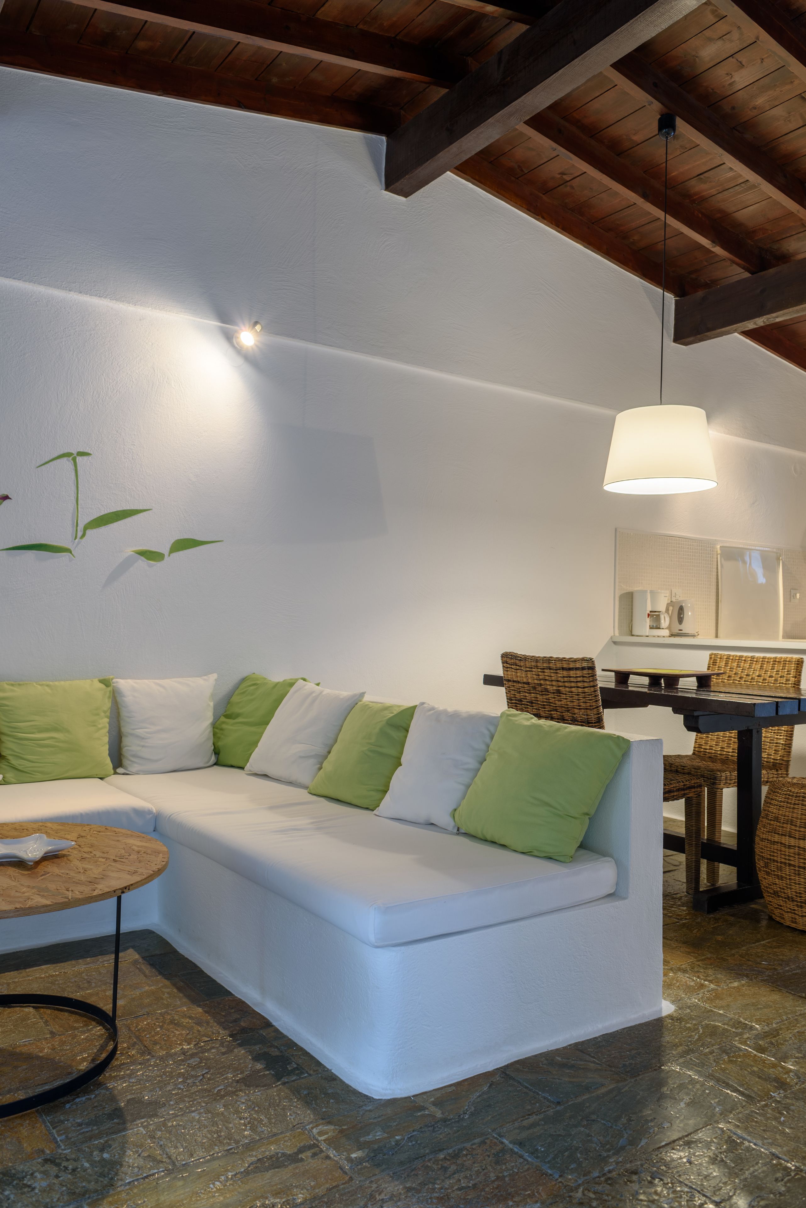 A modern living room with a white sofa, green cushions, a round wooden table, wicker chairs, a hanging light fixture, and a stone-tiled floor.