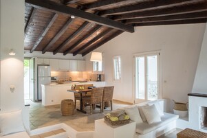 Uma sala de estar ampla com um sofá branco, uma mesa de jantar com cadeiras de vime, uma cozinha com armários brancos e uma lareira.