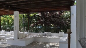 Un patio al aire libre con asientos y mesas blancos acolchados, rodeado de abundante follaje.