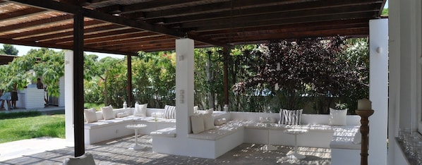 Un patio al aire libre con asientos y mesas blancos acolchados, rodeado de abundante follaje.
