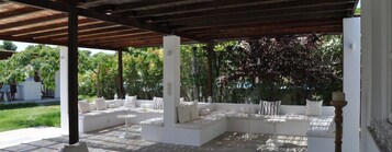 Um pátio externo com assentos e mesas acolchoados em branco, rodeado por vegetação exuberante.