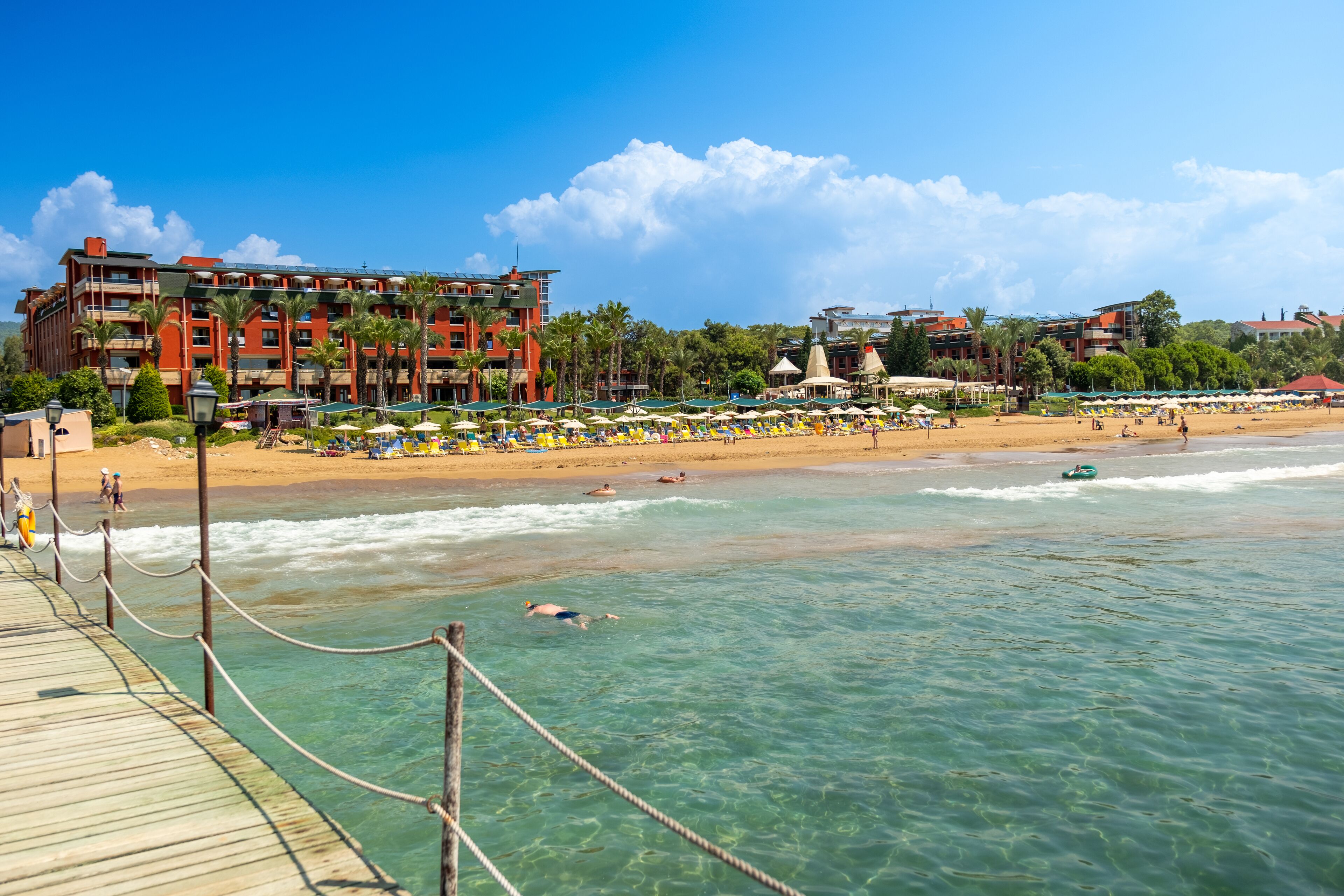 A beach resort with a wooden boardwalk, clear turquoise water, and a sandy beach with umbrellas and people.