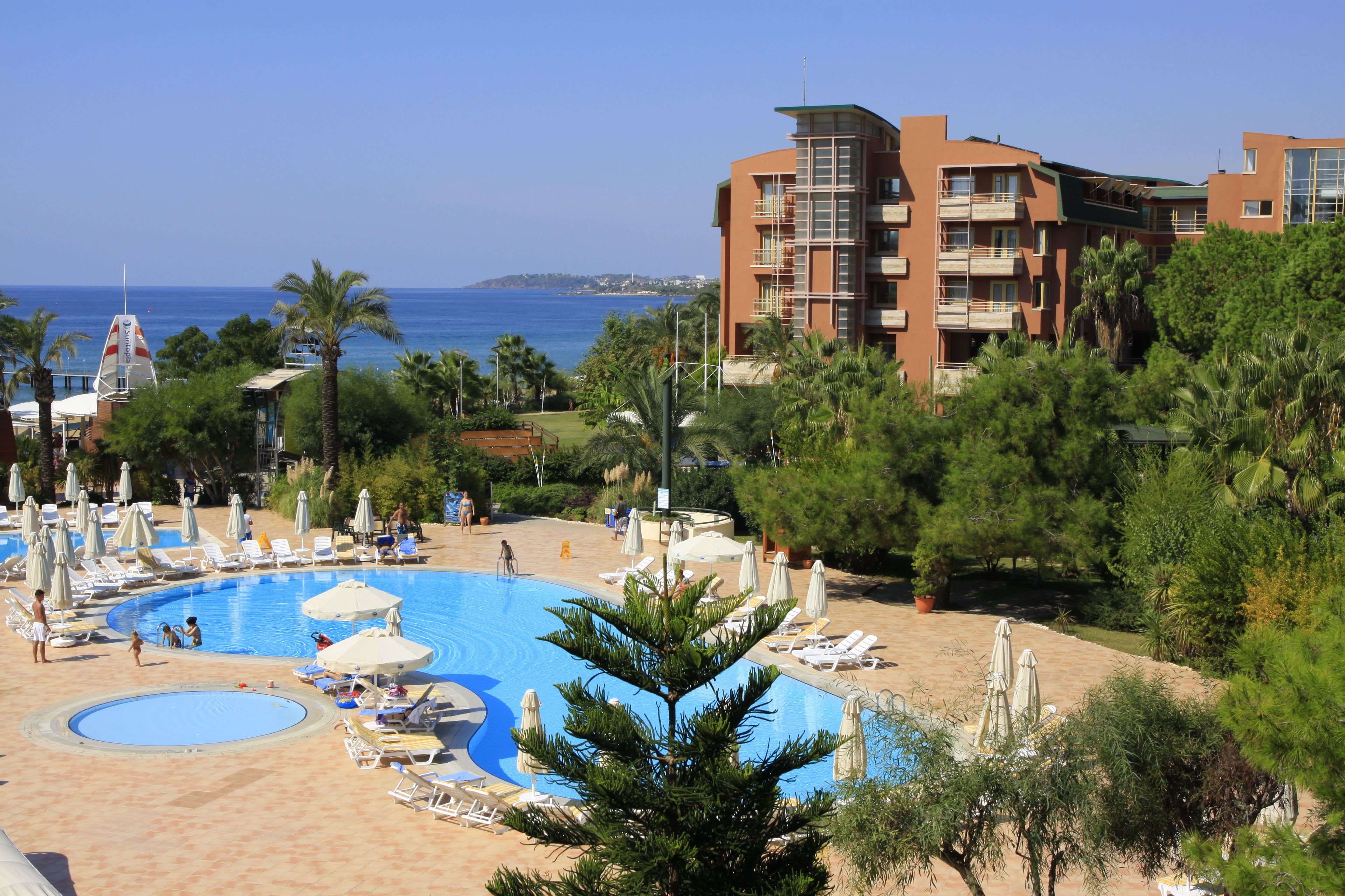 A hotel with a large swimming pool surrounded by lounge chairs and umbrellas, a beach, and a view of the sea.