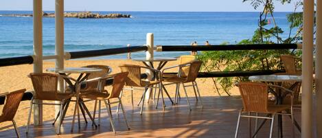 An outdoor seating area with wicker chairs and tables, overlooking a beach and the sea.