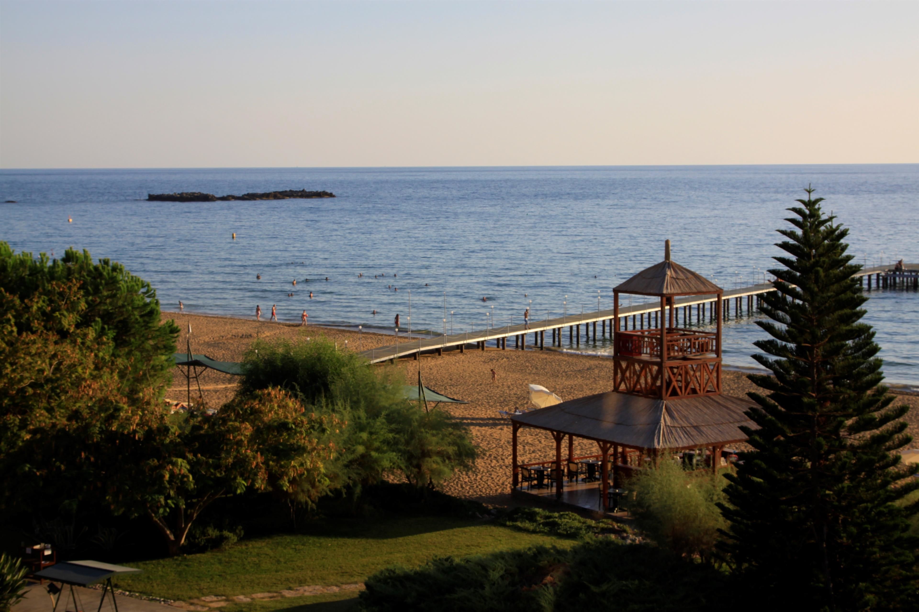 A beach with a wooden gazebo, a pier extending into the water, and a rocky outcrop in the distance.