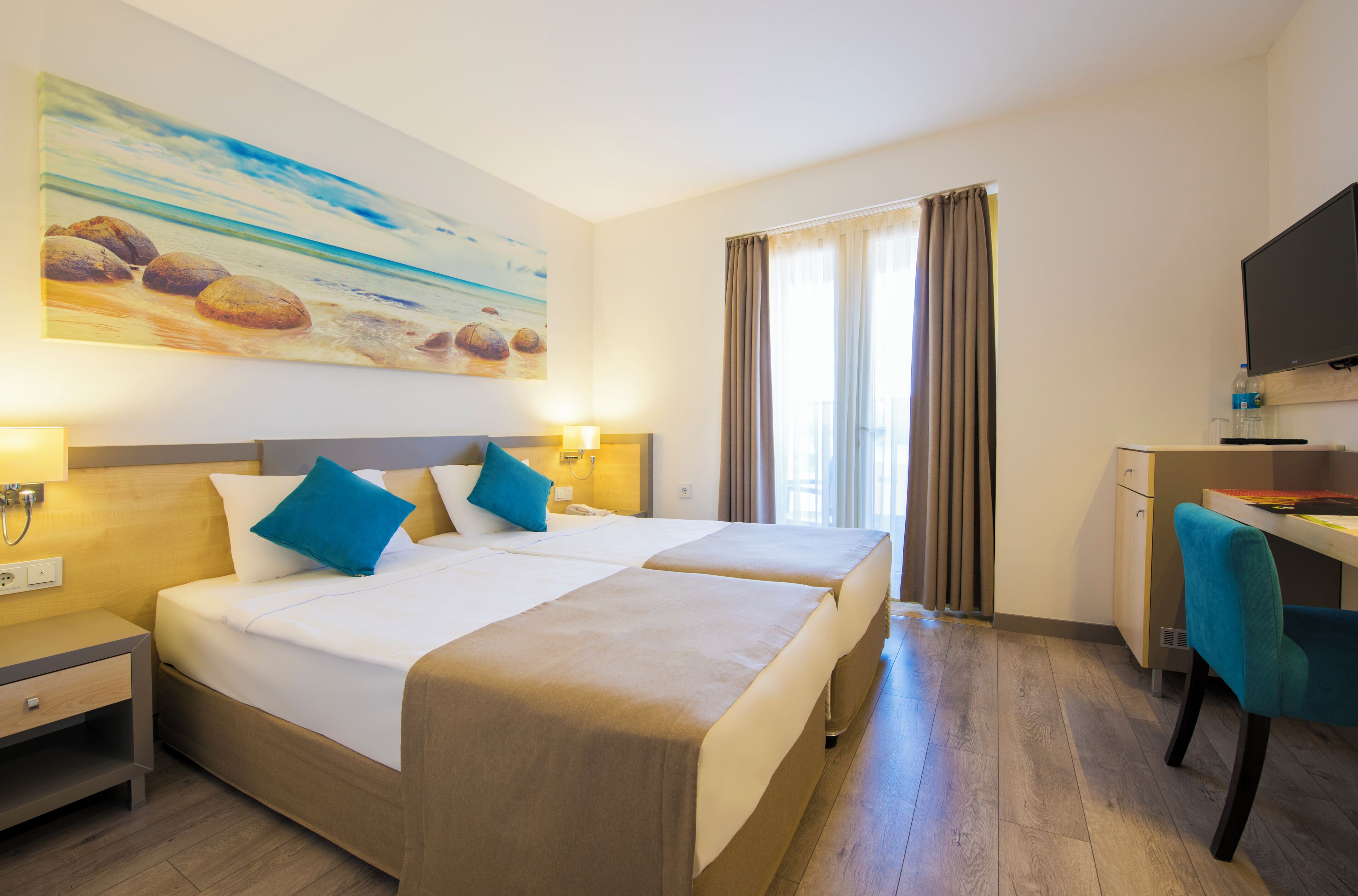 A hotel room with two beds, a desk, a television, and a painting of a beach scene.