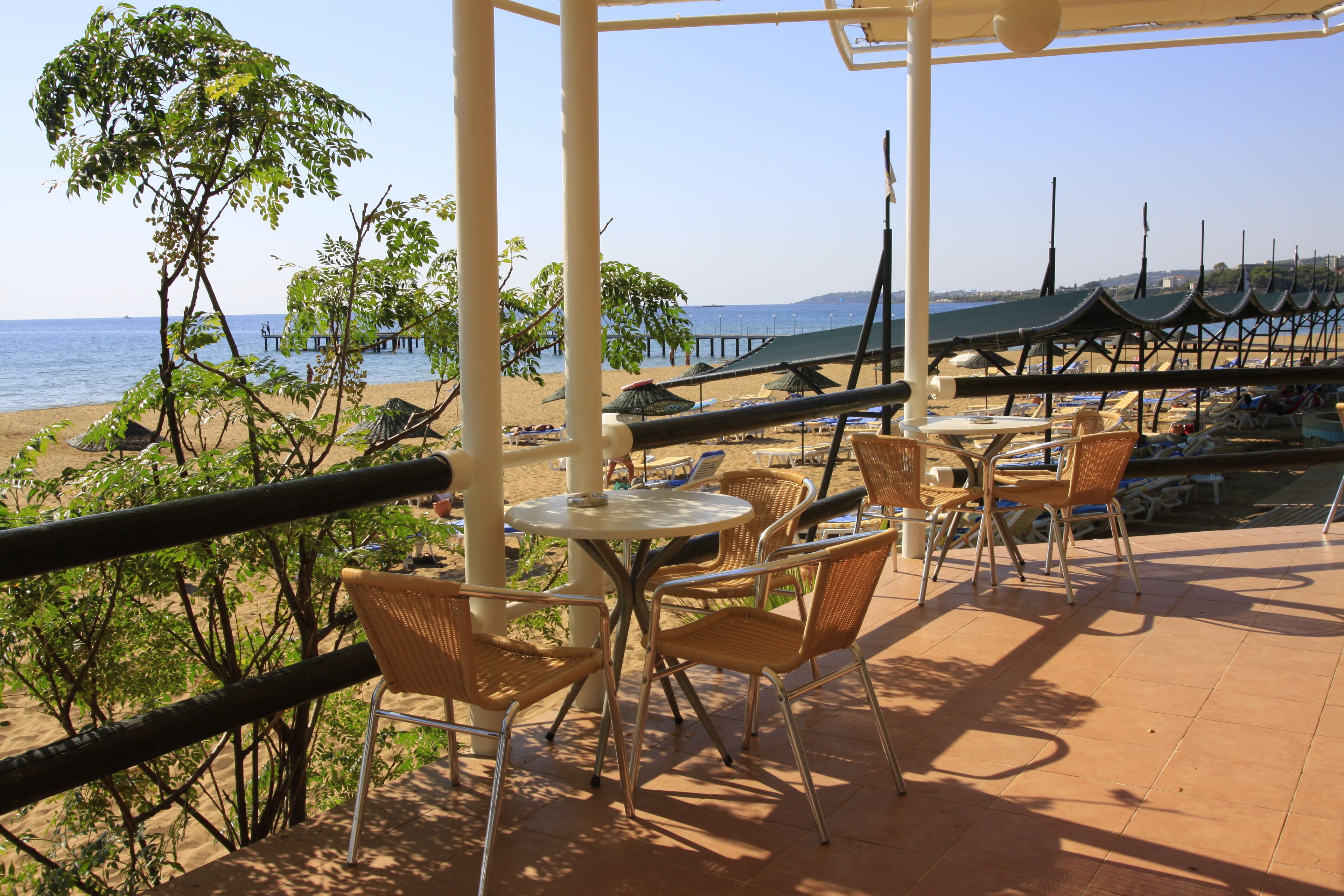 Eine Terrassenförmige Sitzgruppe am Strand mit Tischen und Stühlen, mit Blick auf das Meer und einen Steg.