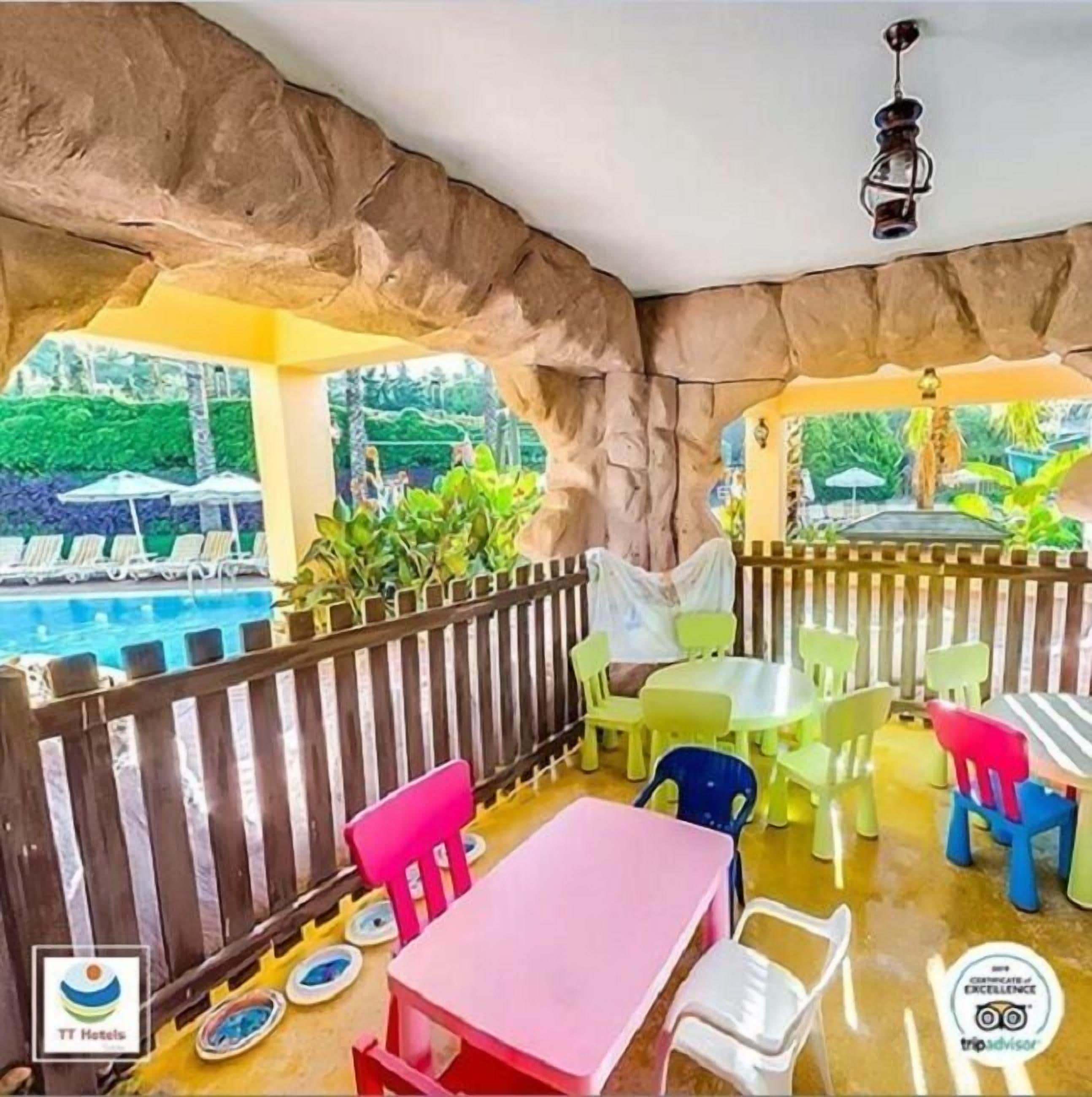 A covered outdoor seating area with colorful plastic chairs and tables, a wooden fence, and a view of a swimming pool.