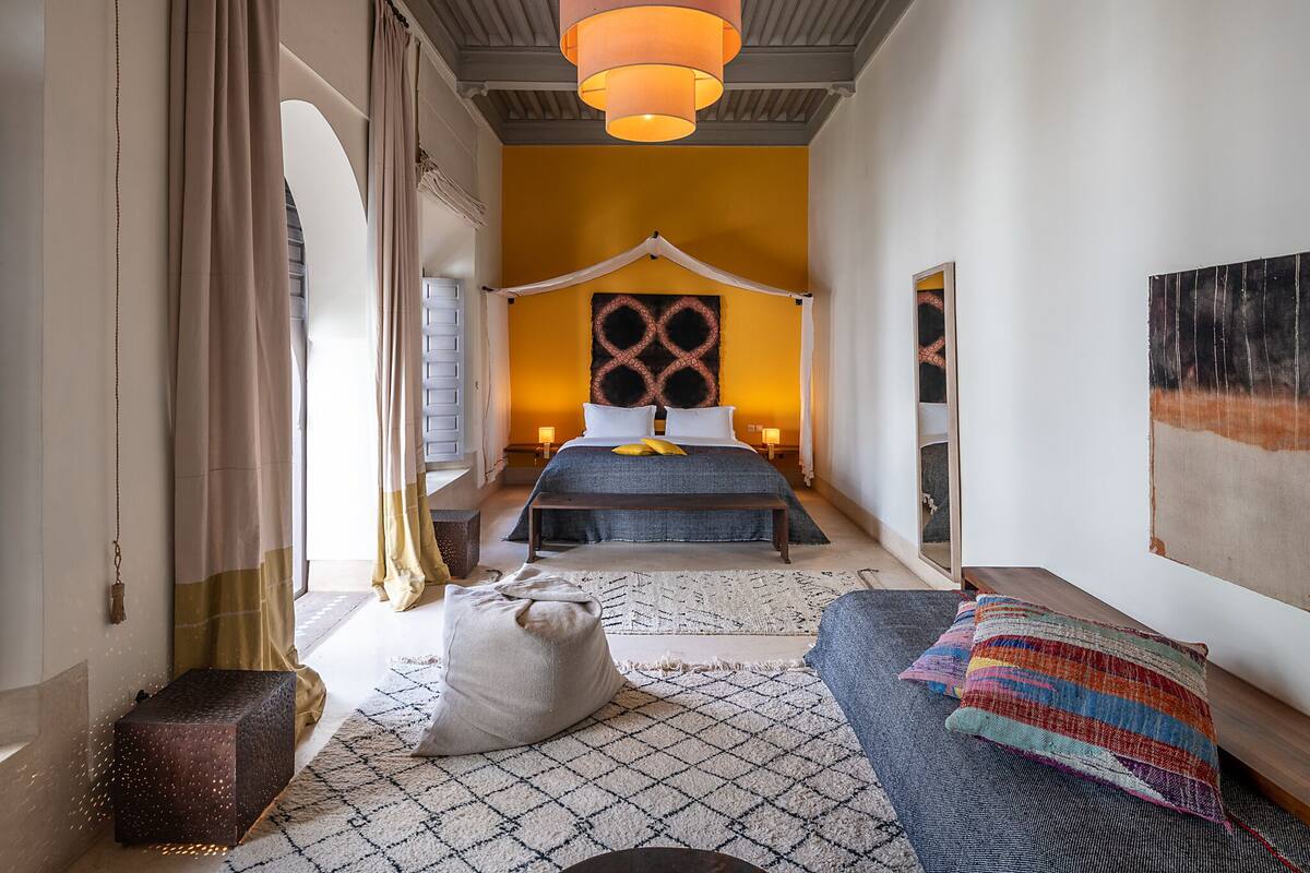 Un dormitorio con cama, mesita de noche, espejo, lámpara colgante y ventana con cortinas.