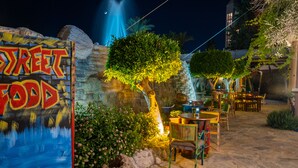 An outdoor dining area with a sign reading "Street Food," tables, chairs, and a waterfall feature.